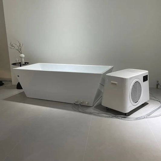 The Lunara Deluxe Plunge, a modern white bathtub, is paired with an advanced cooling portable water chiller on wheels in a minimalist bathroom featuring light gray flooring and a small black side table with a vase and glass.