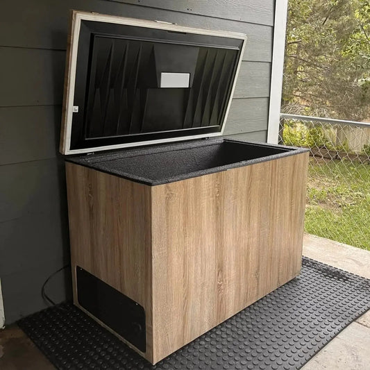 Lunaras The Essential Plunge chest freezer, featuring a wood-like exterior and open black lid, is set on a textured rubber mat on a covered outdoor patio near a chain-link fence and green lawn for cold therapy and wellness.