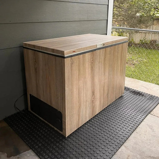 The Essential Plunge by Lunara, a wood-grain chest freezer, sits on a black mat on a covered patio near a gray wall—an ideal cold therapy setup—with a chain-link fence and trees in the background.